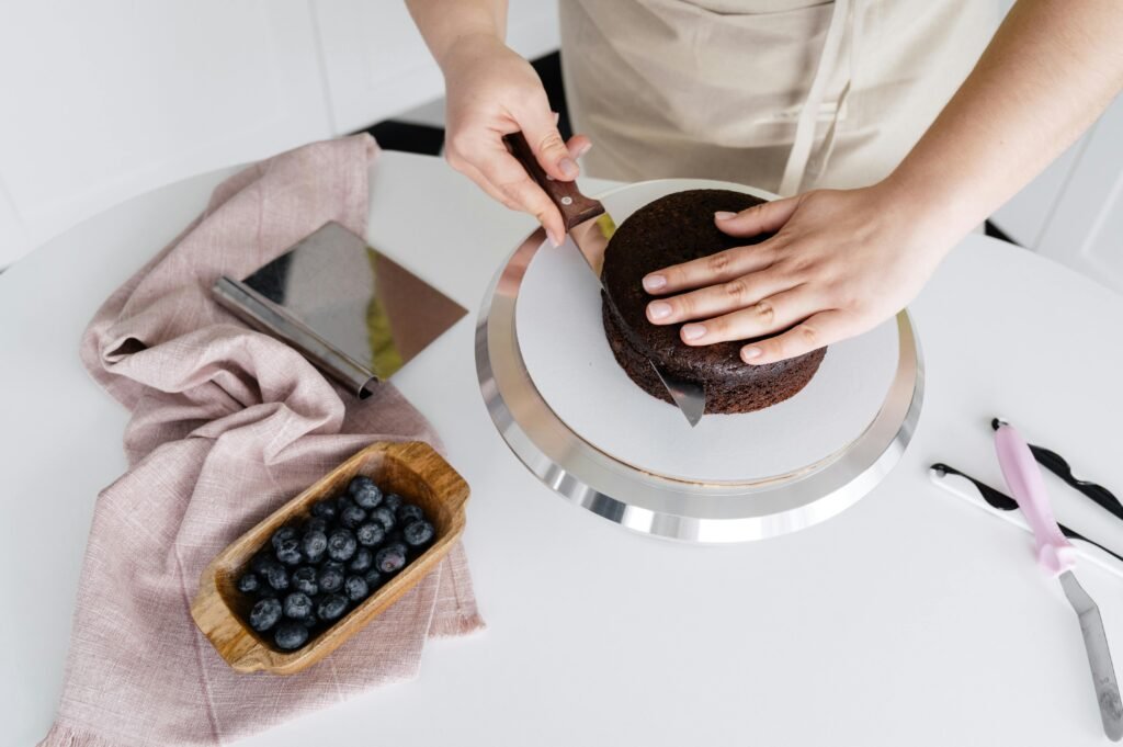 Crop anonymous person cutting delicious homemade cake placed on plate on white table with blueberries and cloth in light kitchen