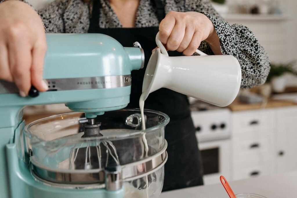 Person in kitchen pouring milk into a stand mixer bowl during baking preparation.